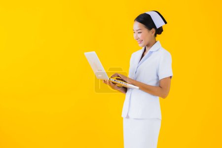 Téléchargez les photos : Portrait belle jeune asiatique femme thai infirmière avec ordinateur portable sur fond jaune isolé - en image libre de droit