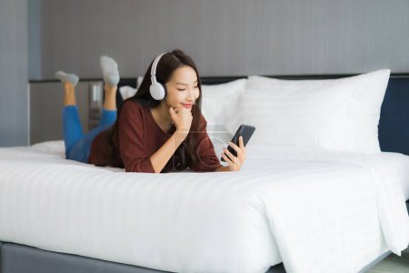 Téléchargez les photos : Portrait belle jeune femme asiatique utiliser téléphone portable intelligent avec casque pour écouter de la musique sur le lit dans la chambre à coucher intérieur - en image libre de droit