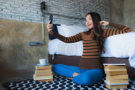 Téléchargez les photos : Jeune femme asiatique en utilisant un téléphone portable intelligent avec livre de lecture et tasse de café dans la chambre à coucher intérieure - en image libre de droit