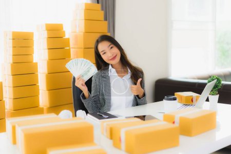 Foto de Retrato hermosa joven mujer asiática trabajo desde casa con portátil en efectivo y caja de cartón listo para el envío de compras de clientes en línea - Imagen libre de derechos
