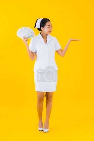 Téléchargez les photos : Portrait belle jeune femme asiatique thai infirmière avec beaucoup d'argent et d'argent et tirelire rose sur fond isolé jaune - en image libre de droit