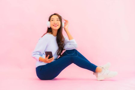 Foto de Retrato hermosa joven asiática mujer uso inteligente teléfono móvil con auriculares para escuchar música sobre fondo rosa - Imagen libre de derechos