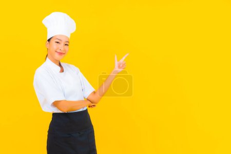 Téléchargez les photos : Portrait belle jeune femme asiatique en chef ou cuisinier uniforme avec chapeau sur fond jaune isolé - en image libre de droit