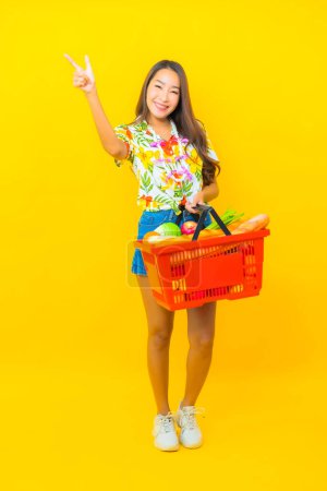 Téléchargez les photos : Portrait belle jeune femme asiatique avec panier d'épicerie du supermarché sur fond jaune - en image libre de droit