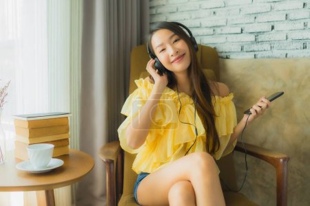 Téléchargez les photos : Portrait jeune femme asiatique assis sur chaise écouter de la musique avec téléphone portable café et livre dans le salon intérieur - en image libre de droit