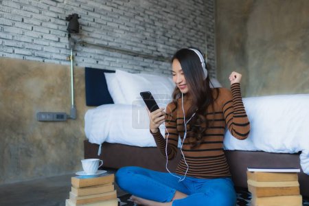 Foto de Mujer asiática joven usando teléfono móvil inteligente con auriculares para escuchar música alrededor de la taza de café y reservar en el interior del dormitorio - Imagen libre de derechos