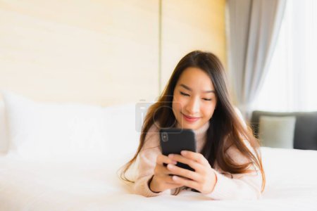 Foto de Retrato hermosa joven mujer asiática con taza de café y teléfono móvil en la cama en el interior del dormitorio - Imagen libre de derechos