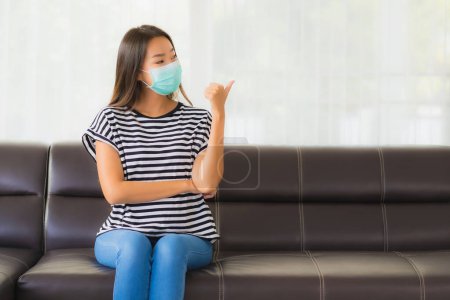 Photo for Portrait beautiful young asian woman with mask stay home on sofa in living room for protect coronavirus or covid19 - Royalty Free Image