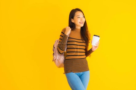 Téléchargez les photos : Portrait belle jeune femme asiatique avec sac à dos et tasse de café dans sa main sur fond jaune isolé - en image libre de droit