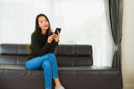 Foto de Retrato hermosa joven asiática mujer uso inteligente teléfono móvil o teléfono celular en sofá en sala de estar interior - Imagen libre de derechos
