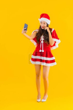 Photo for Portrait beautiful young asian woman wear christmas clothes and hat use smart mobile cell phone on yellow isolated background - Royalty Free Image