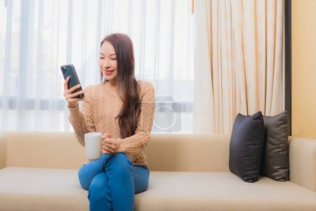 Foto de Retrato hermosa joven mujer asiática relajarse sonrisa feliz con el teléfono inteligente con café en el sofá - Imagen libre de derechos