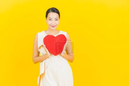 Téléchargez les photos : Portrait belle jeune femme asiatique sourire avec forme d'oreiller de coeur sur fond de couleur jaune - en image libre de droit