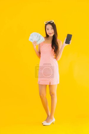 Photo for Portrait beautiful young asian woman show a lot of cash and money with mobile smart phone on yellow isolated background - Royalty Free Image