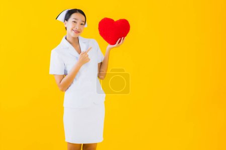 Téléchargez les photos : Portrait belle jeune femme asiatique thai infirmière montrent la forme de l'oreiller cardiaque et le travail à la clinique ou à l'hôpital sur fond jaune isolé - en image libre de droit