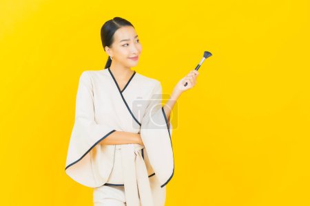 Téléchargez les photos : Portrait belle jeune femme d'affaires asiatique avec maquillage pinceau cosmétique sur fond jaune - en image libre de droit