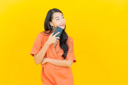 Téléchargez les photos : Portrait belle jeune femme asiatique sourire avec téléphone portable intelligent sur fond de couleur jaune - en image libre de droit