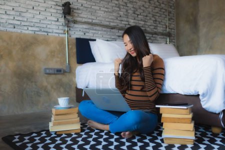 Foto de Mujer asiática joven usando computadora portátil con taza de café y libro en el interior del dormitorio - Imagen libre de derechos
