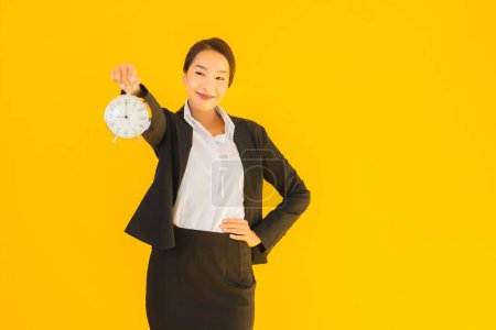 Téléchargez les photos : Portrait belle jeune femme asiatique montrer l'heure de l'horloge ou de l'alarme sur fond jaune isolé - en image libre de droit