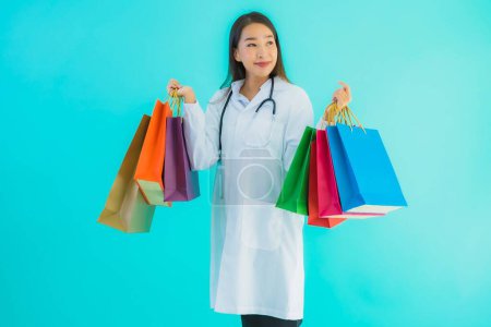 Téléchargez les photos : Portrait belle jeune femme médecin asiatique avec sac à provisions coloré du centre commercial sur fond bleu isolé - en image libre de droit