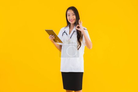 Téléchargez les photos : Portrait belle jeune asiatique médecin femme utiliser appareil tablette intelligente sur fond jaune isolé - en image libre de droit
