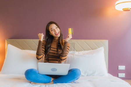 Téléchargez les photos : Portrait belle jeune femme asiatique utiliser ordinateur portable sur le lit avec carte de crédit à l'intérieur de la chambre - en image libre de droit