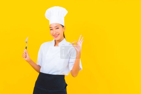 Téléchargez les photos : Portrait belle jeune femme asiatique porter chef ou cuisinier uniforme avec cuillère et fourchette sur fond jaune isolé - en image libre de droit