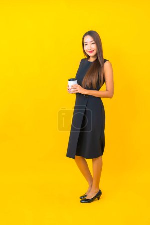 Foto de Retrato hermosa joven mujer de negocios asiática con taza de café y teléfono móvil inteligente sobre fondo amarillo - Imagen libre de derechos