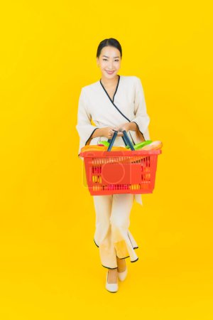Téléchargez les photos : Portrait belle jeune femme asiatique d'affaires avec panier épicerie de supermarché sur fond jaune - en image libre de droit