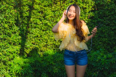 Téléchargez les photos : Jeune femme asiatique utilisant un téléphone portable intelligent avec casque pour écouter de la musique autour du jardin extérieur vue sur la nature - en image libre de droit
