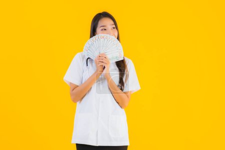 Foto de Retrato hermosa joven asiática médico mujer con un montón de dinero en efectivo y dinero en amarillo aislado fondo - Imagen libre de derechos