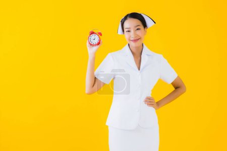 Téléchargez les photos : Portrait belle jeune asiatique femme thai infirmière montrer horloge ou alarme sur fond jaune isolé - en image libre de droit