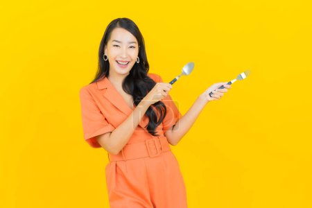 Téléchargez les photos : Portrait belle jeune femme asiatique sourire avec cuillère et fourchette sur fond de couleur jaune - en image libre de droit