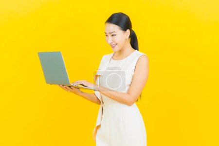 Téléchargez les photos : Portrait belle jeune femme asiatique sourire avec ordinateur portable sur fond jaune isolé - en image libre de droit