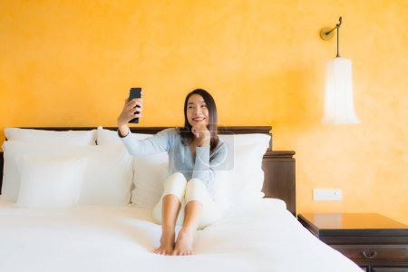 Foto de Retrato hermosa joven mujer asiática utilizando el teléfono celular móvil en la cama con taza de café en el interior del dormitorio - Imagen libre de derechos