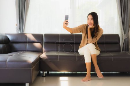 Téléchargez les photos : Portrait belle jeune femme asiatique utiliser smartphone mobile ou téléphone portable avec tasse de café sur canapé salon intérieur - en image libre de droit