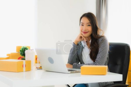 Foto de Retrato hermosa joven mujer de negocios asiática trabajo desde casa con teléfono móvil portátil con caja de cartón listo para el envío de compras en línea - Imagen libre de derechos