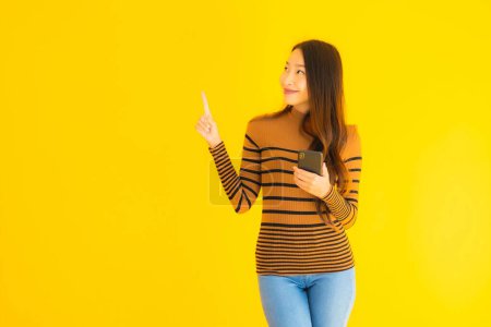 Téléchargez les photos : Belle jeune femme asiatique utiliser téléphone mobile intelligent ou téléphone portable avec beaucoup d'action sur fond jaune isolé - en image libre de droit