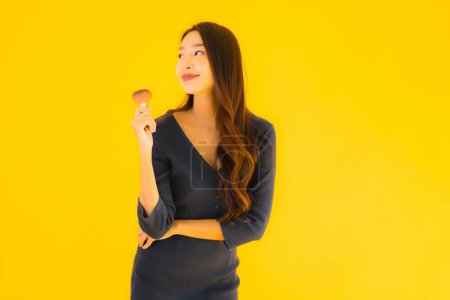 Téléchargez les photos : Portrait belle jeune femme asiatique avec pinceau de maquillage sur fond jaune isolé - en image libre de droit