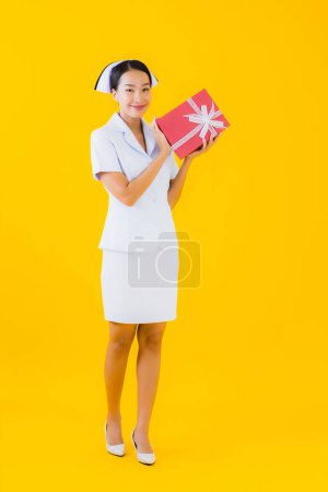 Foto de Retrato hermosa joven asiática mujer tailandesa enfermera show rojo regalo caja en amarillo aislado fondo - Imagen libre de derechos