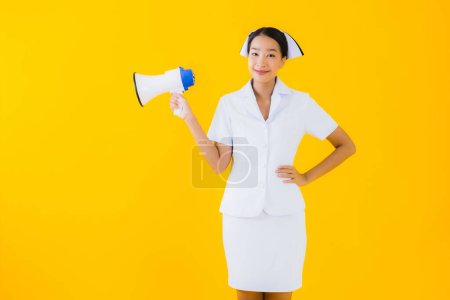 Téléchargez les photos : Portrait belle jeune asiatique femme thai infirmière avec mégaphone pour communiquer en clinique et à l'hôpital sur fond jaune isolé - en image libre de droit