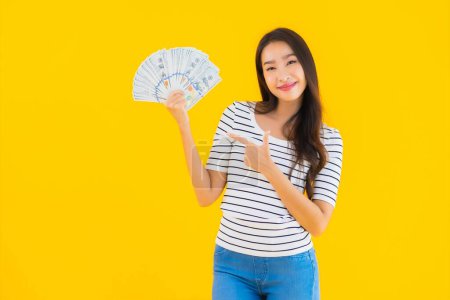 Foto de Retrato hermosa joven asiática mujer mostrar un montón de dólares en efectivo o dinero en amarillo aislado fondo - Imagen libre de derechos