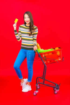 Téléchargez les photos : Portrait belle jeune femme asiatique avec panier d'épicerie du supermarché sur fond rouge isolé - en image libre de droit