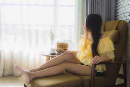 Téléchargez les photos : Portrait jeune asiatique femme assis sur canapé chaise et lire livre avec tasse de café dans salon intérieur - en image libre de droit