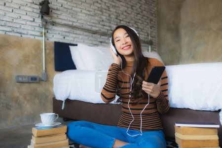 Photo for Young asian woman using smart mobile phone with headphone for listen music around coffee cup and book in bedroom interior - Royalty Free Image