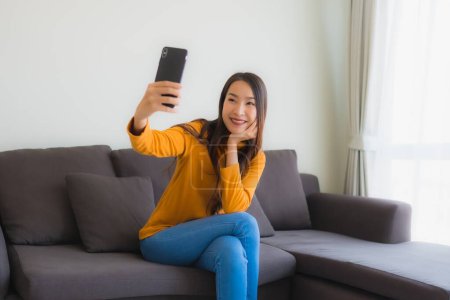 Téléchargez les photos : Portrait jeune femme asiatique utilisant téléphone portable intelligent sur canapé avec oreiller dans le salon intérieur - en image libre de droit