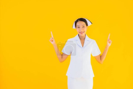Téléchargez les photos : Portrait belle jeune infirmière asiatique thai dans beaucoup de travail d'action pour le patient en clinique et à l'hôpital sur fond jaune isolé - en image libre de droit