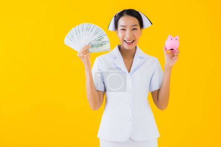 Foto de Retrato hermosa joven asiática mujer tailandesa enfermera con un montón de efectivo y dinero y rosa alcancía en amarillo aislado fondo - Imagen libre de derechos