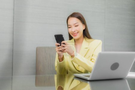 Téléchargez les photos : Portrait belle jeune femme asiatique utiliser ordinateur portable avec téléphone portable intelligent sur la table de travail à l'intérieur - en image libre de droit