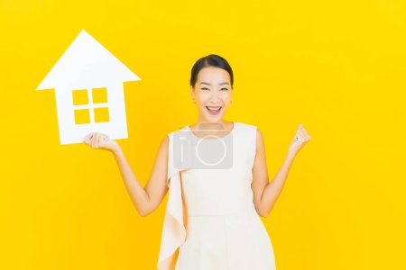 Téléchargez les photos : Portrait belle jeune femme asiatique avec maison ou maison signe de papier sur fond de couleur jaune - en image libre de droit